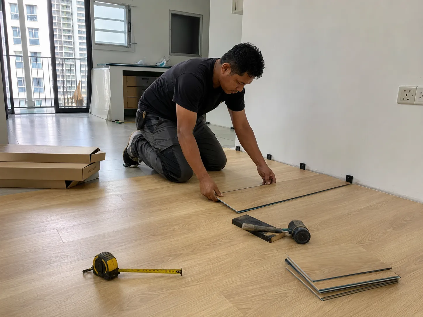 安装师傅在 Condo 单位内铺设锁扣式 vinyl 板材,墙边放有定位垫片、橡胶槌,旁边还有几箱板材